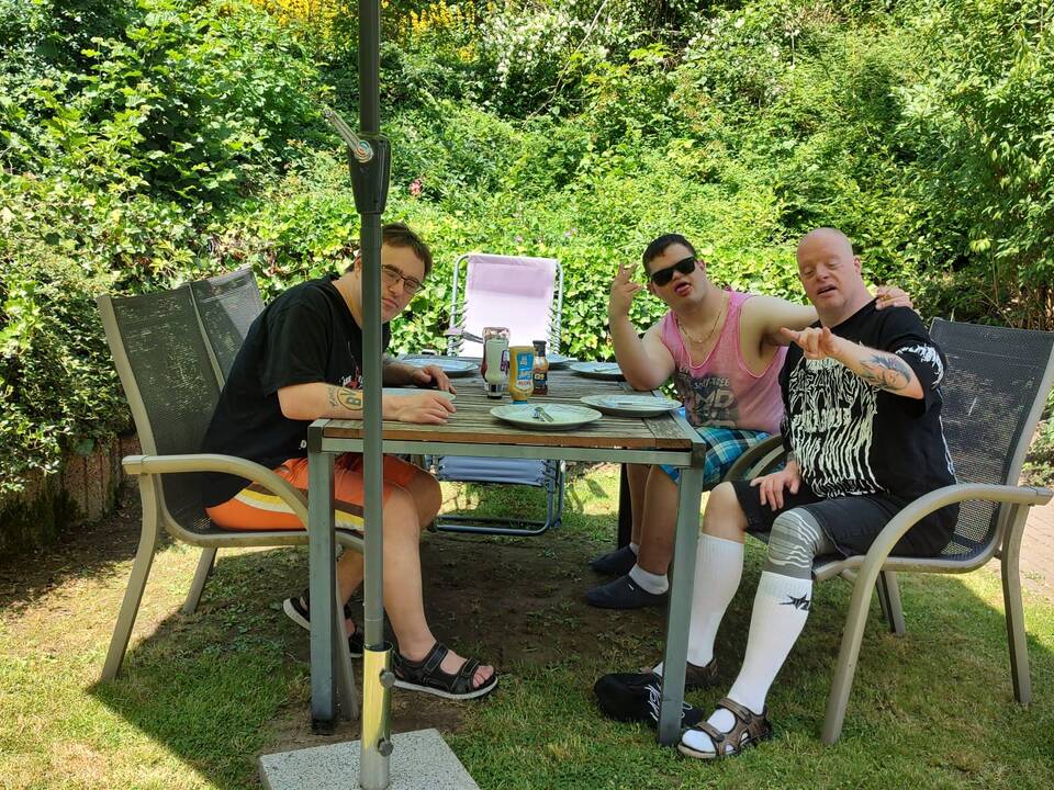 Drei Männer sitzen unterm Sonnenschirm am Tisch im Garten.