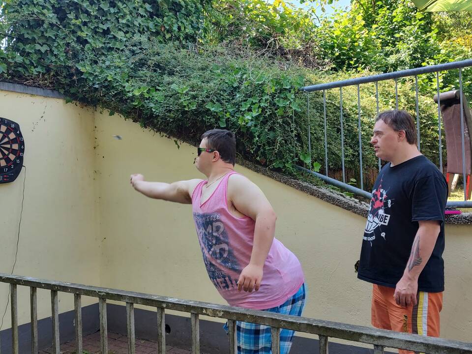 Zwei Männer spielen Dart, einer hat eine Sonnenbrille an.