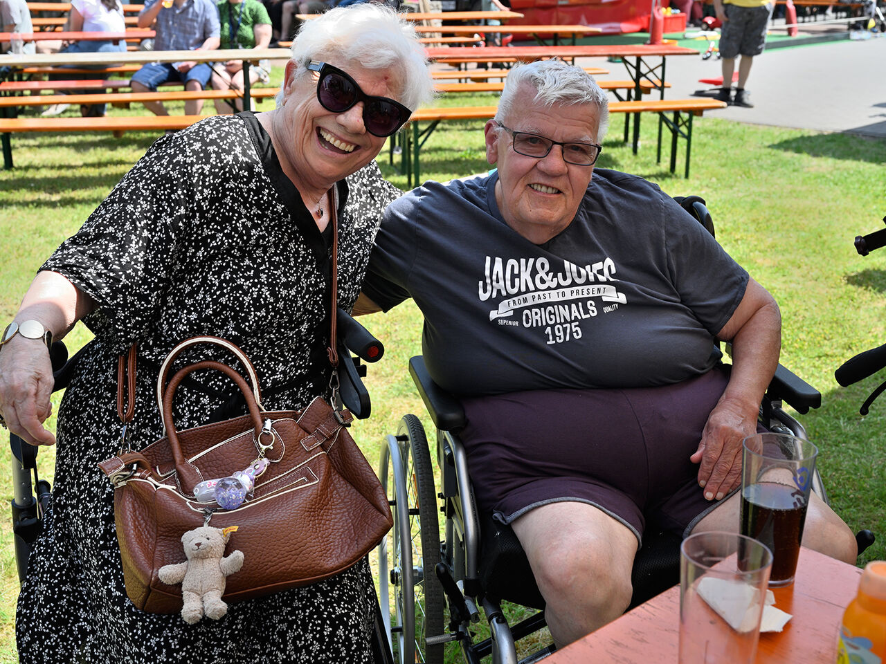 EIn Mann und eine Frau haben sich im Arm, der eine im Rollstuhl die Frau sitzt auf einem Rollator.