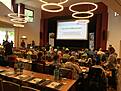 Viele Menschen sitzen an Tischen in einem großen Saal.