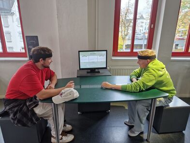 Zwei junge Männer haben ein Band am Kopf, sitzen an einem Tisch und schauen auf einen Monitor.