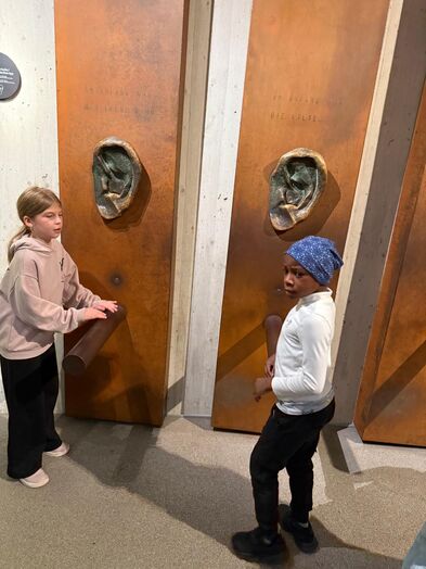 Zwei Kinder stehen vor einer Wand mit zwei großen Ohren.