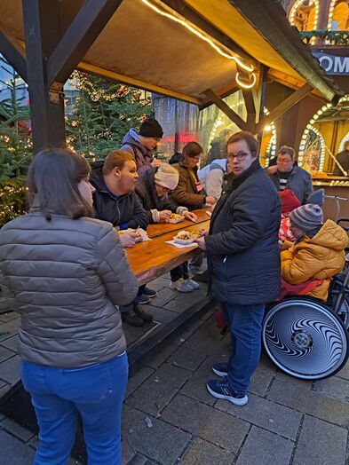 Menschen mit und ohne Hilfsmittel stehen mit Essen an einem großen Stehtisch der überdacht ist.