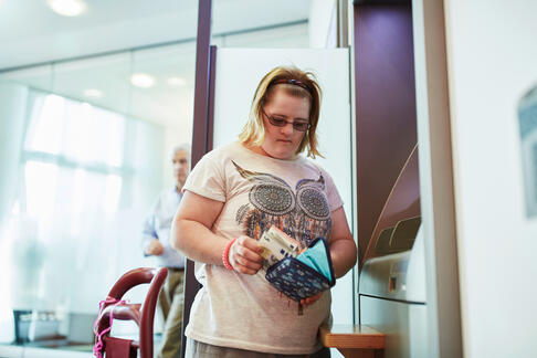Das Bild zeigt eine Frau die Geld am Bankautomaten abgehoben hat und in ihre Geldbörse steckt