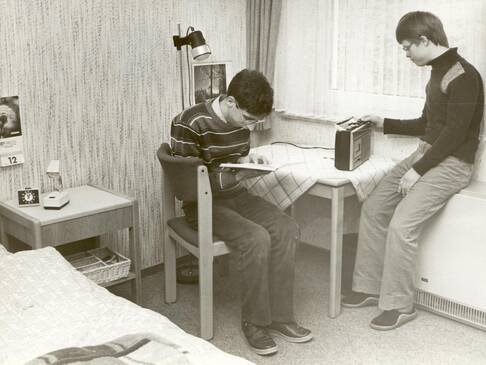 Zwei Personen sitzen an einem Tisch. Einer schaut in ein Buch der andere schaut ein Radio an.