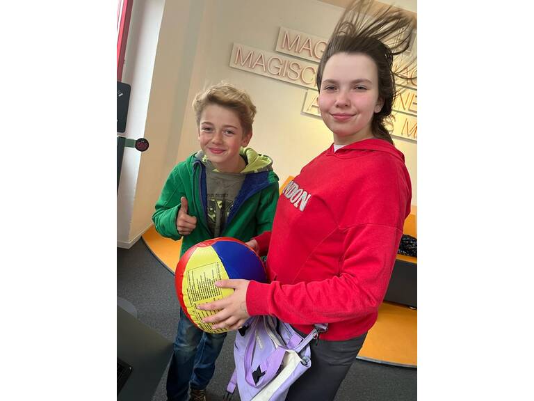 Zwei Jungendliche stehen in einem Windstrom und das eine Kind hält einen Ball in der Hand.