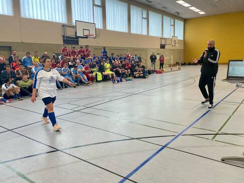 Eine Fussballerin geht durch die Halle im Hintergrund viele Fussballer die auf Bänken sitzen.
