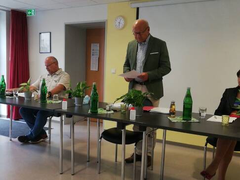 Ein Mann steht hinter dem Tisch, zwei Personen sitzen.