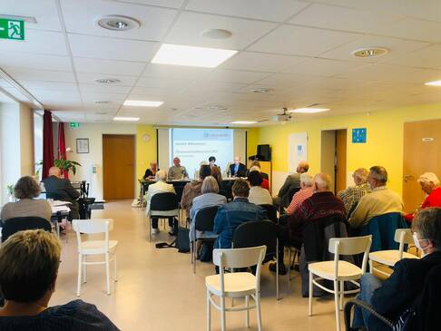 Viele Menschen sitzen in einem Raum und folgen einer Präsentation an der Wand
