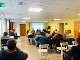 Viele Menschen sitzen in einem Raum und folgen einer Präsentation an der Wand