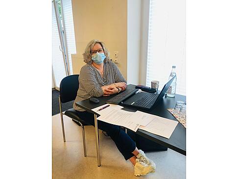 Eine Frau sitzt mit Maske vor einem Laptop am Tisch.