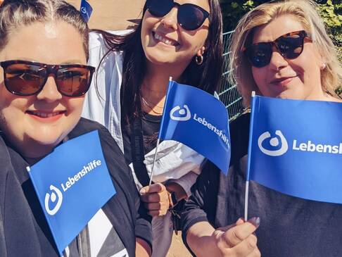 Drei Frauen mit Sonnenbrille und Lebenshilfefahnen stehen in der Sonne.