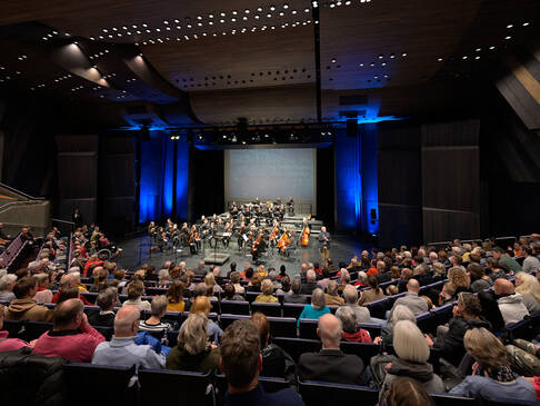 Eine Saalaufnahme, mit Puplikum und einem Orchester auf der Bühne.