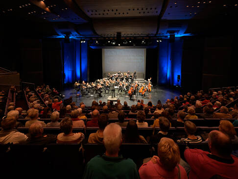 Eine Saalaufnahme, mit Puplikum und einem Orchester auf der Bühne.
