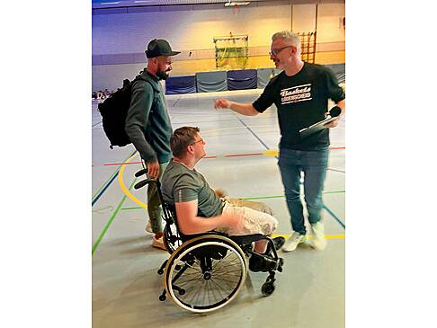 Ein Rollstuhlfahrer, ein Mann mit Käppi steht daneben und ein anderer Mann zeigt auf diesen Mann.