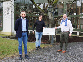 Drei Männer vor einem Gebäude, zwei halten einen Scheck in der Hand