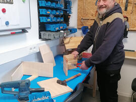 Mann steht an einer Werkbank und hat Holz in der Hand.