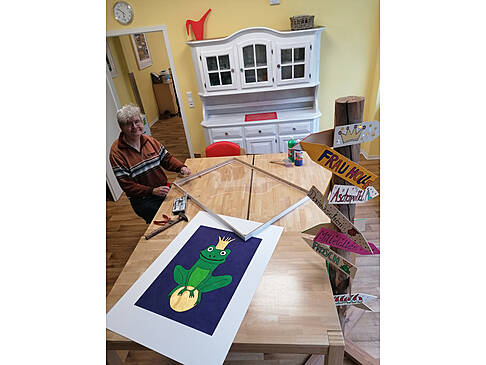 Mann sitzt am Tisch mit Werkzeug in der Hand, auf dem Tisch liegt ein Bild mit einem Froschkönig und ein Rahmen.