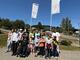 Eine Gruppe aus Kindern und jngen Menschen mit und ohne Rollstuhl stehen für ein Gruppenfoto auf einem großen Platz zusammen. Im Hintergrund sind zwei Flaggen mit der Aufschrift "Campingpark im Bergisches Land" zu sehen.