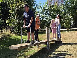 Eine Gruppe von Kindern und Erwachsenen balanciert auf einer Holzwippe im Freien. Sie üben bei Sonnenschein und umgeben von grünen Bäumen ihre Geschicklichkeit.