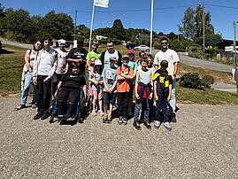 Eine Gruppe aus Kindern und jngen Menschen mit und ohne Rollstuhl stehen für ein Gruppenfoto auf einem großen Platz zusammen. Im Hintergrund sind zwei Flaggen mit der Aufschrift "Campingpark im Bergisches Land" zu sehen.