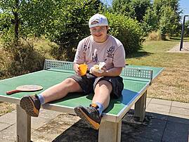 Eine junge Person macht eine Pause auf einer Tischtennisplatte im Freien und genießt einen Snack. Die Person lächelt zufrieden in die Kamera.