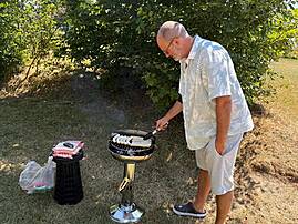 Ein Mann grillt bei sonnigem Wetter Würstchen auf einem kleinen Säulengrill. Er steht auf einer Wiese und wendet konzentriert das Grillgut.