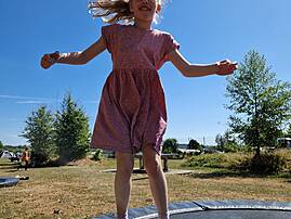 Ein junges Mädchen schwebt beim Trampolinspringen in der Luft. Ihre Haare wehen und sie strahlt vor Freude unter dem blauen Himmel.