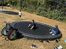 Zwei Kinder sitzen auf einem in den Boden eingelassenen Trampolin. Daneben im Gras steht ein Rollstuhl mit bunten Speichen.