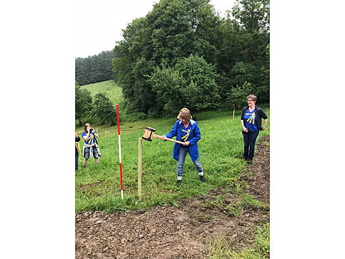 Eine Person schlägt ein Rundholz in den Boden auf einer Wiese.