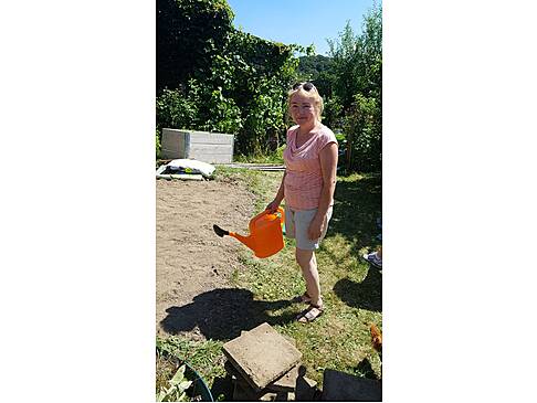Eine Frau steht im Garten mit einer orangen Gieskanne in der Hand.