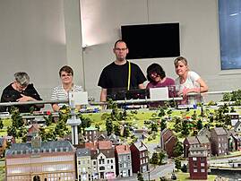 Mehrere Menschen stehen hinter einer großen Modelleisenbahn mit detailreicher Stadtlandschaft, Häusern, Straßen, Bäumen und Zügen. Im Hintergrund sind ein Fernseher und eine helle Innenwand zu sehen.