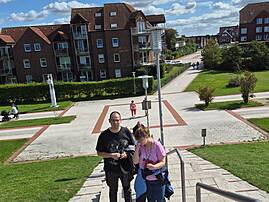 Zwei Menschen gehen bei sonnigem Wetter eine breite Treppe hinauf. Im Hintergrund stehen Wohnhäuser, gepflegte Grünflächen und weitere Menschen auf einem Weg.