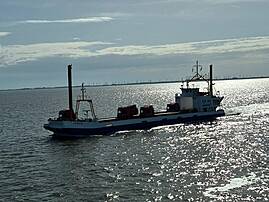 Ein Frachtschiff mit mehreren Lastwagen fährt über das glitzernde Meer. Am Horizont sind Windräder und ein weiter, bewölkter Himmel zu sehen.