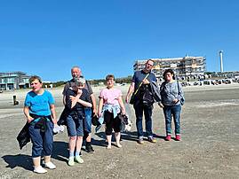 Eine Gruppe aus sechs Menschen steht bei sonnigem Wetter an einem kiesigen Strand. Im Hintergrund sind Gebäude, Strandkörbe und ein weiter blauer Himmel zu sehen.