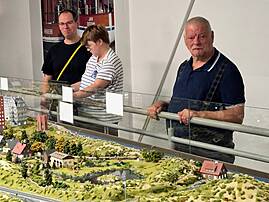 Drei Menschen betrachten hinter einer Glasscheibe eine detailreiche Modelllandschaft mit Straßen, Häusern, Bäumen und kleinen Figuren. Im Hintergrund hängt ein großes Bild mit Schiffen.