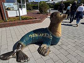 Auf einem Platz steht eine große, bunt bemalte Seehundfigur mit der Aufschrift „NORDDEICH.“ Im Hintergrund sind ein Hinweisschild zur Seehundstation und mehrere Menschen zu sehen.