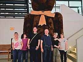 Sechs Menschen stehen in einem Gebäude vor einer großen sitzenden Bärenfigur mit einer karierten Schleife. Im Hintergrund sind Treppen, ein Hinweis mit der Aufschrift „Berlin“ und ein heller Innenraum zu sehen.