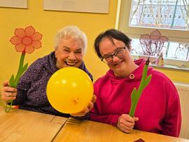 Zu sehen sind zwei Frauen sitzend am Tisch und lachen, sie halten einen gelben Luftballon in der Mitte und jede hält eine gebastelte Blume in der Hand