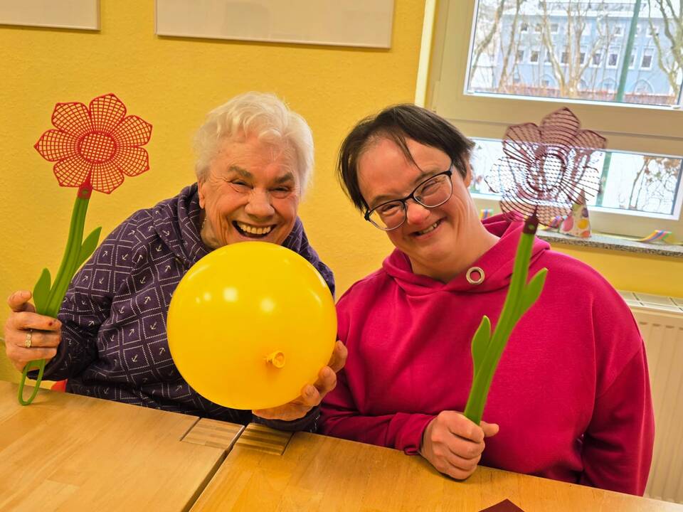 Zu sehen sind zwei Frauen sitzend am Tisch und lachen, sie halten einen gelben Luftballon in der Mitte und jede hält eine gebastelte Blume in der Hand