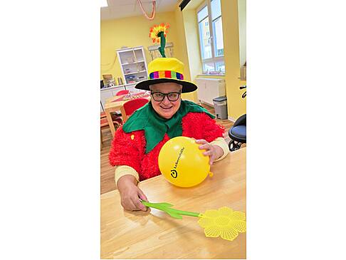 eine Frau sitzt im rot-grünen Blütenkostüm am Tisch mit gelben Hut, auf dem Tisch hält sie einen gelben Luftballon in der Hand und in der anderen Hand eine gelbe Plastikblume am Stiel
