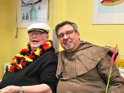 auf dem Bild sieht man zwei Männer mit Karnevalschmuck, der eine mit einer Deutschland-Hulakette und der andere in einem Priesterkostüm