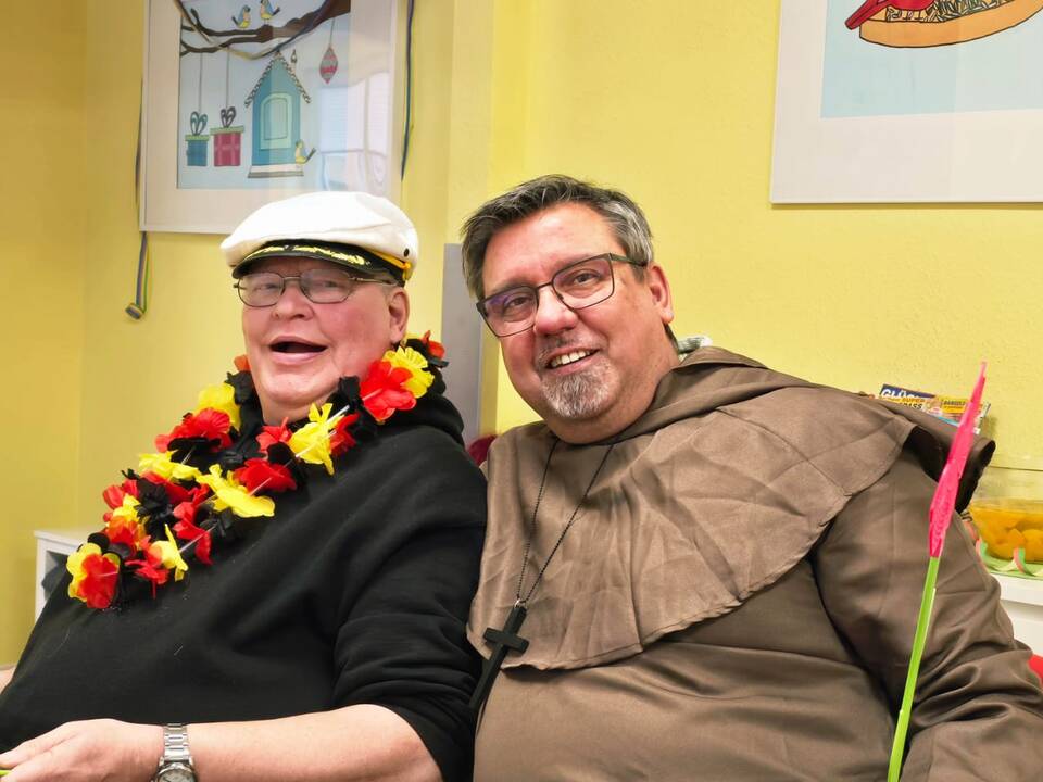 auf dem Bild sieht man zwei Männer mit Karnevalschmuck, der eine mit einer Deutschland-Hulakette und der andere in einem Priesterkostüm