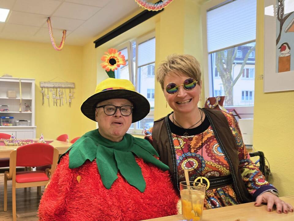auf dem Bild sieht man zwei Frauen am Tisch in Karnevalskostümen, die eine Frau trägt ein Blütenkostüm mit gelben Hut auf dem Kopf und die ander Frau ist als Hippie verkleidet, vor ihnen steht eine Getränk im Glas mit Strohhalm