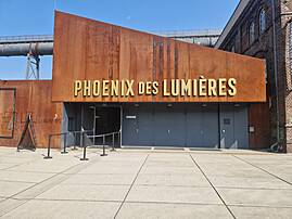Das Bild zeigt den Eingangsbereich des "Phoenix des Lumières" in Dortmund. Das moderne Gebäude hat eine rostige Fassade aus Cortenstahl, auf der in großen, goldenen Buchstaben der Name der Ausstellung steht.