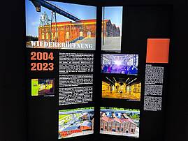 Eine Informationstafel mit dem Titel "Wiedereröffnung" und den Jahreszahlen 2004 und 2023 beleuchtet die Geschichte eines Ortes. Mehrere Fotos und Texte dokumentieren die Verwandlung eines alten Industriegebäudes in einen modernen Kulturort.