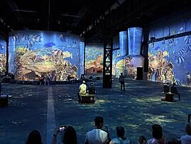 Menschen sitzen in einer großen Halle und blicken auf eine riesige Projektion, die eine Meeresschildkröte und viele Fische zeigt. Die immersive Ausstellung lässt die Besucherinnen und Besucher in eine digitale Unterwasserwelt eintauchen.