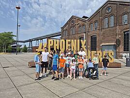 Eine Gruppe von Jugendlichen und Erwachsenen posiert für ein Gruppenfoto vor dem Haupteingang des "Phoenix des Lumières". Im Hintergrund ist das Backsteingebäude und eine alte Industrieanlage zu sehen.