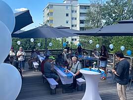 Eine Gruppe von Menschen feiert auf einer sonnigen Holzterrasse. Sie sitzen an langen Tischen unter großen Sonnenschirmen und unterhalten sich in entspannter Atmosphäre.