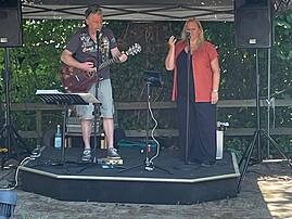 Ein Musiker mit Akustikgitarre und eine Sängerin stehen auf einer kleinen Bühne im Freien. Sie treten unter einem Pavillon auf und sorgen für die musikalische Unterhaltung der Gäste.